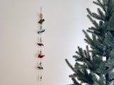 A vibrant felt American bird garland hangs vertically against a plain white wall, creating a serene, nature-inspired display. This handmade felt garland features intricately crafted figures of a colorful hummingbird, blue jay, cardinal, and chickadee, interspersed with small green felt leaves along a thin string. To the right, a section of a green Christmas tree adds a touch of seasonal warmth to the scene.