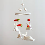 A felt baby mobile featuring bunnies and carrots is suspended against a plain white background. White felt balls at the top connect via strings to a felt ring. From the ring hang three fluffy white felt bunnies with pink noses and black eyes, alongside two vibrant red-orange felt carrots with green leafy tops.