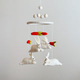 A charming felt baby mobile, featuring white bunnies and bright orange carrots, is suspended against a light gray background. The whimsical design showcases detailed bunnies with pink noses and stitched eyes, appearing to interact with carrots topped with green felt. Above the central felt ring, white felt balls and delicate strings support this playful and imaginative mobile, ideal for a nursery.