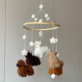 A charming felt baby mobile, featuring Highland Cow figures, hangs against a plain grey wall, creating a soothing atmosphere. It has a light brown wooden frame with a cluster of white felt balls at the top. Four fluffy felt Highland cows in caramel, dark brown, light brown, and white, along with white felt stars and clouds, are suspended by white threads. The mobile's palette of white, beige, caramel, brown, and chocolate offers a soft, inviting appearance.