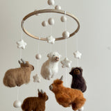 A felt baby mobile featuring Highland Cows is displayed against a plain white background, creating a calm and playful atmosphere. It features a wooden ring from which four felt Highland cow figures hang at varying lengths on soft cotton threads. The cows are crafted with intricate needlework, showcasing soft textures and fluffy fur accents in shades of white, beige, caramel, brown, and chocolate. This delightful and creative design is ideal for a nursery.