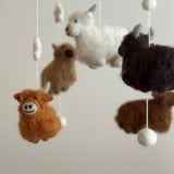 A felt baby mobile featuring charming Highland cows is suspended from a wooden frame against an off-white background. Five felted cows, in white, caramel-brown, and dark brown, hang with white felt balls and spindle shapes. Each cow displays fluffy fur, small gray horns, and intricate needlework, adding a playful touch.