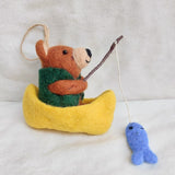 A charming felt bear fishing ornament is poised on a textured off-white background. This unique Christmas tree ornament features a brown bear in a bright yellow tiny boat, wearing a dark green vest. The bear holds a brown fishing rod with a white string and a small blue fish. A jute string loop is attached for hanging.