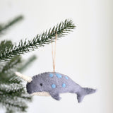 A handmade felt blue narwhal ornament is delicately hung on a Christmas tree branch, set against a bright, festive background. This unique wool felt narwhal ornament features a soft gray body with playful blue spots and a white belly. It has black eyes and a distinctive stitched ivory tusk, with a jute string attached for hanging.