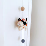 A charming 3D needle-felted cat from a Felt Cat Garland hangs against a bright white door. Crafted from 100% wool, the white felt cat features distinct black, orange, and white markings, small pink ears, a stitched pink mouth, and matching pink paws. It is suspended by a black string with a tan felt ball above and a gray one below, emphasizing its whimsical, handcrafted design.