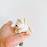 A charming light beige 3D wool felt cat figure, part of a felt cat garland, is held in a person's palm against a bright white background. The cat has a small tail, stubby legs, and black embroidered facial features. A grey felt ball from the garland is visible below the cat.