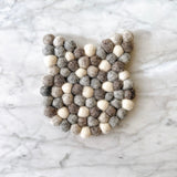 One charming wool felt cat coaster from a set of four rests on a sleek marble backdrop. This handcrafted, eco-friendly felt cat coaster features subtle cat ears and is made from tightly packed wool felt balls in shades of white, gray, and brown, creating a textured appearance.