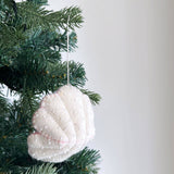 A white wool felt seashell ornament, part of a sea animal felt ornament set, is gracefully suspended from a green Christmas tree branch. This handcrafted felt ornament features a textured surface embellished with tiny white beads. It hangs amidst green pine needles and brown pinecones, evoking a simple, warm, and inviting holiday decor.