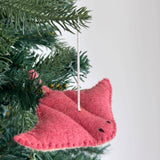 A coral felt stingray ornament, part of a sea animals felt Christmas ornament set, hangs on a lush green Christmas tree. This handmade wool felt ornament features a wide, flat body with a segmented pattern, small black stitched eyes, and a top loop for hanging.
