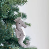 A seahorse wool felt ornament, part of a Sea Animals felt Christmas ornament set, hangs on a festive Christmas tree. It is crafted from textured grey wool felt with a lighter white chest and a simple black dot for an eye, bringing a joyful, rustic charm to the holiday setting. A thin cotton string is attached for hanging.