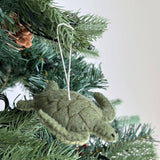 A green turtle felt Christmas ornament, part of a sea animal set, hangs from a pine needle branch of a Christmas tree. Crafted from soft wool felt, this festive ornament features a segmented shell and small, detailed flippers, adding a handcrafted touch.