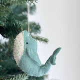 A wool felt whale Christmas ornament hangs gracefully from a Christmas tree's evergreen branches. This charming felt ornament features a soft, textured light turquoise body and a creamy white belly, with contrasting green thread stitching. A white cotton string is attached, adding a touch of rustic elegance.