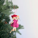 A charming handmade felt Little Sister mouse Christmas ornament hangs gracefully from a green Christmas tree. This delightful needle felted mouse, with a cream-colored body, wears a vibrant pink dress and a jaunty red hat. A red cotton string is attached, ready for hanging on the festive boughs.