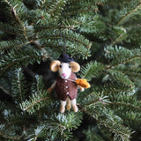 A charming wool felt mouse Christmas ornament hangs from a lush green Christmas tree branch. This 3D felt mouse ornament features tan inner ears, a pink nose, and a black hat. It wears a brown vest with three white buttons and holds a yellow corncob, adding to its whimsical design.