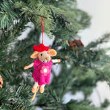 A charming wool felt mouse Christmas ornament, representing the 'Little Sister' from a set, hangs from a Christmas tree branch adorned with pine needles and warm white lights. The handmade mouse is needle-felted, dressed in a bright pink dress with a white flower detail and a small red hat, suspended by a delicate red cotton string.