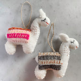 Two white felt llama Christmas ornaments are presented against a plain background, showcasing their handcrafted charm. Each unique Christmas ornament is adorned with a detailed blanket and a jute string for hanging. One llama features an orange, pink, and white striped blanket with delicate stitching, while the other displays a brown and grey blanket. Subtle closed eyes and a small mouth give these felt llama ornaments a gentle, soft appearance.