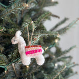 A handcrafted felt llama Christmas ornament hangs on a warmly lit Christmas tree. This charming white llama ornament features a colorful saddle in pink, brown, and yellow, with delicate stitched eyes and a red mouth. It is suspended by a jute string, blending into the festive, blurred background.