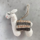 A handcrafted white wool felt llama Christmas ornament, part of a set, rests on a gray marble surface, showcasing its charm. The llama features a multi-striped blanket in brown, gray, and beige with red and white stitched accents. It has stitched closed eyes and a red stitched mouth, completed by a jute string for hanging, embodying a rustic, warm aesthetic.