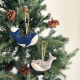 Two felt whale Christmas ornaments are suspended on a festive Christmas tree adorned with pinecones, creating a warm holiday scene. One whale is deep blue with white accents and small white dots, while the other is light gray with white accents. Both handmade felt ornaments feature a charming, handcrafted appearance.