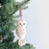 A hand-stitched felt owl Christmas ornament hangs gracefully from a green pine branch on a festive Christmas tree. The charming felt owl features a light beige, plump body and a round head. It has small, dark eyes and delicate stitched details suggesting feathers, complete with a red and white cotton string for hanging.