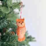 A hand-stitched felt owl ornament, part of a Felt Christmas Ornaments Set, hangs festively on a green Christmas tree branch against a blurred white background. The charming owl ornament is crafted from orange and brown felt, featuring a tan face, small ears, and delicate white stitches on its body. It is suspended by a red and white cotton string.