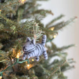 A gray tabby felt cat Christmas ornament hangs on a festive Christmas tree illuminated with warm white lights. This charming handcrafted felt ornament features a round body with distinctive black stripes and a matching striped tail. It has perky ears and an embroidered face with eyes, a nose, and a mouth. A jute string is attached for hanging.