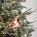A charming handmade felt cat ornament, part of a Felt Christmas Ornaments set, hangs gracefully from a festive Christmas tree adorned with string lights. This unique felted cat ornament is light brown with darker brown stripes, featuring a tail and ears. A jute string is attached for hanging.