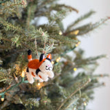 A festive felt cat ornament hangs on a lush green Christmas tree, its branch illuminated by warm white lights. Crafted from felt, this unique Christmas ornament features a playful cat in white, orange, and black with small, perky ears. A jute string allows this charming handmade felt cat ornament to hang gracefully, adding a cozy touch to holiday decor.