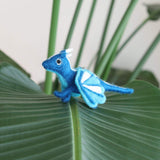A handmade blue felt dragon Christmas ornament is gracefully displayed on a textured leaf, set against vibrant green plant leaves and a white wall. This captivating flying dragon ornament is predominantly blue with white accents on its wings and head. It features a soft, wool-like texture, small black eyes, and a metal loop at the top for hanging, adding a playful charm to holiday decor.