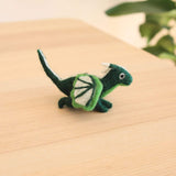 A green felt flying dragon Christmas ornament rests on a light wooden surface, illuminated by soft lighting with a plant subtly in the background. This intricately-crafted felted dragon features white accents, including detailed wings with green trim, a white-tipped tail, and small white horns. A green cotton string is attached, ready for hanging.