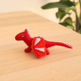 A vibrant red felt flying dragon ornament rests on a wooden surface with a blurred green background. This playful handmade felt Christmas ornament features intricate white accents on its tail and wings, along with small white horns.