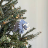 A handmade felt triceratops Christmas ornament hangs on a Christmas tree with warm lights in a festive, blurred background. The felt triceratops ornament has a light purple body, three white horns, small black eyes, and red nostrils. Its frill is also light purple, and a jute string is attached for hanging.