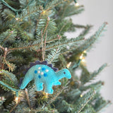 A charming felt Christmas dinosaur ornament is displayed on a vibrant Christmas tree adorned with warm, glowing lights. This light blue felt dinosaur features darker blue accents along its spine and playful white and blue polka dots on its side. A jute string is attached for easy hanging, blending with the cozy holiday atmosphere.