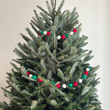 A festive Felt Christmas Pompom Ball Garland is draped around the branches of a Christmas tree, creating a playful holiday atmosphere. The garland features spherical felt pompom balls in red, green, and white, evenly spaced on a string.