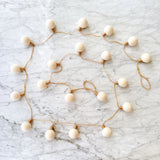 A wool felt lightbulb garland rests on a light gray marble surface, viewed from above. This felt color ball lightbulb garland features spherical, off-white wool felt balls that mimic lightbulbs. Each 'lightbulb' is secured with a thin, rustic brown string, creating a minimalist and appealing design with neutral tones.