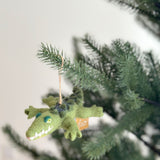 A felted wool gator ornament, designed to appear crawling, hangs from a vibrant green Christmas tree. This unique Christmas tree ornament features a textured body with meticulously crafted details like its eyes, teeth, and back. A jute string allows the handcrafted felt gator to hang gracefully.
