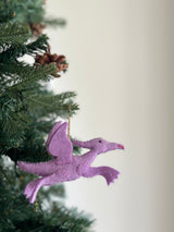A vibrant purple pterosaur felt dinosaur ornament is suspended by a jute string on a Christmas tree, nestled among green needles and pine cones. This wool felt ornament appears to be in mid-flight, showcasing hand-stitched edges, small black eyes, and a red beak. Its whimsical design adds a charming touch to the festive decor.