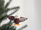 A felt eagle ornament hangs on a green Christmas tree branch, suspended by a jute string against a plain white background. This patriotic felt eagle ornament features a white head and tail, a brown body, and wings detailed with white star patterns. Bright orange feet and a beak add a pop of color, enhancing its American spirit-inspired design.
