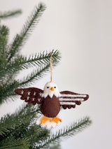 A felt eagle ornament hangs from a Christmas tree branch, creating a festive and patriotic atmosphere. This needle felted eagle ornament features a white head, yellow beak, brown body, outstretched brown wings with white stitches, and orange felt talons, adding handcrafted charm.