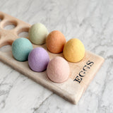 Six handcrafted felt Easter eggs are nestled on a bright marble surface, creating a soft and cheerful springtime mood. These spherical wool felt ornaments display a gentle texture in pastel shades of green, orange, yellow, pink, and purple.