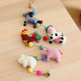 A whimsical felt farm animal garland is displayed on a light wood surface. This handmade wool felt garland features five 3D needle-felted farm animals: a gray horse, a vibrant red and yellow rooster, a white and black milk cow, a pink pig, and a white sheep. Colorful felt balls adorn the string connecting each animal, with a small copper bell hanging at one end.