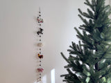 A felt farmland animal garland hangs on a white wall beside a green Christmas tree, creating a festive atmosphere. This 4-foot handmade wool felt garland features five detailed 3D needle-felted animals: a black and white milk cow, a grey horse, a white sheep, a red rooster, and a pink pig. Grey felt balls separate the animals along the string, which ends with a copper bell.