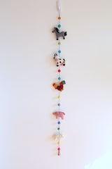 A felt farmland animal garland hangs playfully against a plain white wall, featuring five 3D felted figures. The decorative garland includes a gray horse, a black and white milk cow, a red and yellow rooster, a pink pig, and a white sheep, with a copper bell at its end.