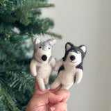 A handcrafted Alaskan Husky Dog felt finger puppet is held by a female hand, set against a blurred Christmas pine tree backdrop. This fair-trade wool felt puppet features a gray and white color scheme, erect triangular ears, and a rounded snout, with intricate needle felt detailing, presented playfully.