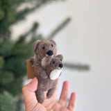 A handmade felt American River Otter finger puppet, featuring a light brown mother otter cradling a smaller white baby otter with a brown face, is held by a fair-skinned hand. Both otters have black eyes and noses. This unique handcrafted felt puppet is set against a blurred Christmas pine tree and a light gray wall.