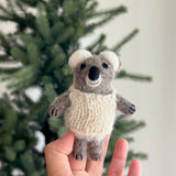 A cozy, handmade wool felt baby koala finger puppet is gently held by a female hand, set against a blurred, warm background suggesting a Christmas tree. This adorable koala puppet, crafted from soft gray wool felt, wears a white woven sweater and features small round white ears, a black nose, and tiny black claws. This unique, handcrafted felt toy is lovingly made in Nepal, reflecting its fair-trade origin and artisanal quality.