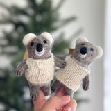 A meticulously handcrafted wool felt baby koala finger puppet is gently held in a hand, displayed against a softly blurred festive Christmas tree backdrop, evoking a playful and inviting mood. The charming gray koala puppet features soft gray fur, fluffy white ears, small black eyes, and a stitched smile, all adorned with a cozy hand-knitted white sweater.