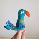 A handcrafted wool felt blue parrot finger puppet is showcased on a fingertip against a plain background. The vibrant bird features a rounded head, an orange beak, and layered blue plumage with hints of yellow. This unique, needle-felted toy, handmade in Nepal, highlights detailed texture.