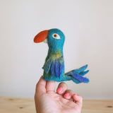 A handmade needle-felted blue parrot finger puppet, crafted in Nepal, is held upright by a female hand with pink nails, against a light background with a light wood surface. The 3D parrot puppet features vibrant blue plumage, a blend of yellow and green on its chest, darker blue accents near its wings and tail, a prominent orange beak, and a small white eye.