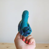 A handcrafted, needle felted blue parrot finger puppet, made from 100% wool, is poised as if in flight against a neutral backdrop on a light wood surface. This vibrant kids finger puppet features a rounded 3D body and textured blue wings with color variations. Its stylized design with minimal facial features is ideal for interactive finger play.